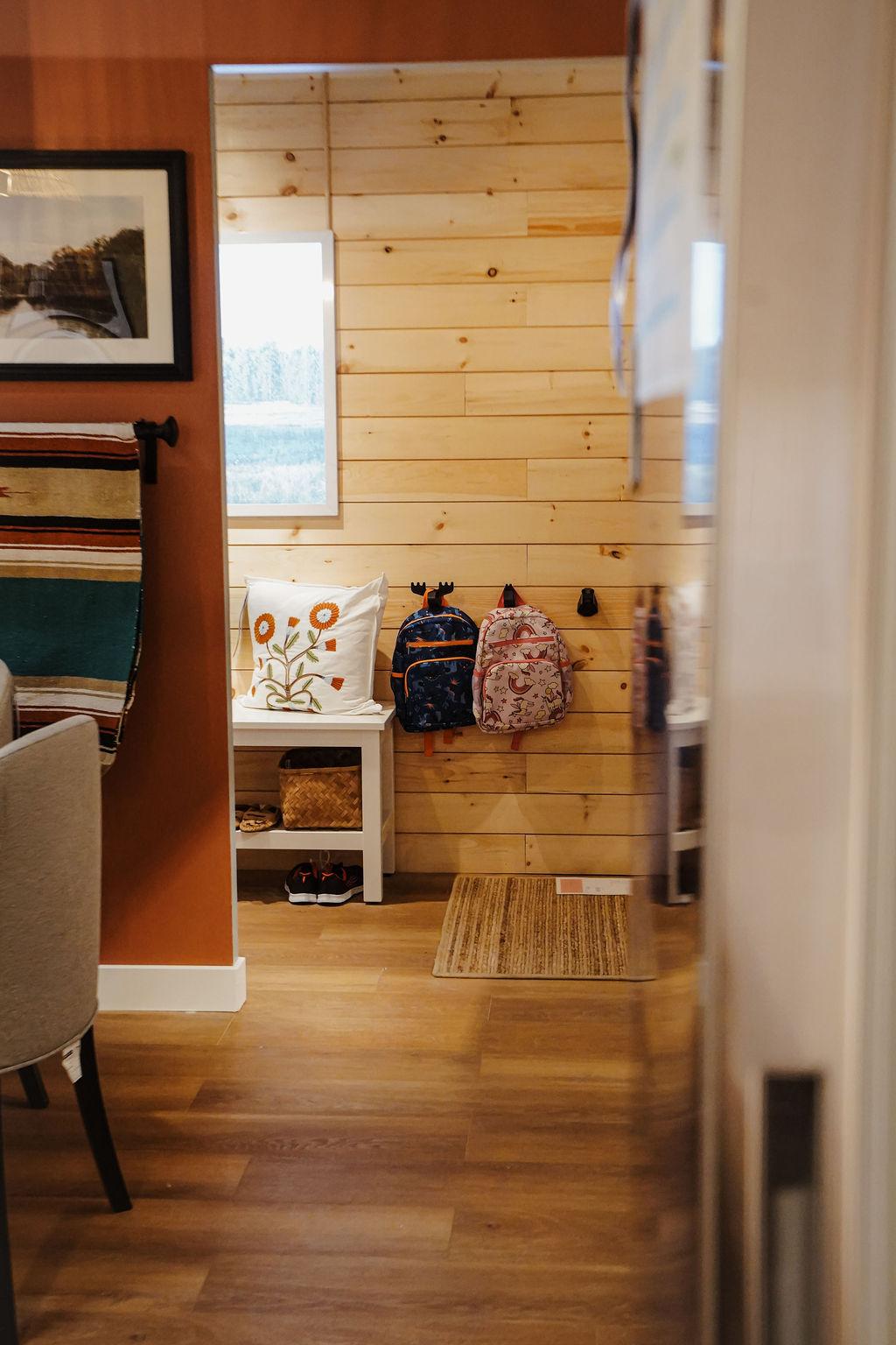 Plan large d’un montage-décor montrant l’arrière d’un mur et des lattes de bois, avec des sacs à dos pour enfants suspendus à des crochets.