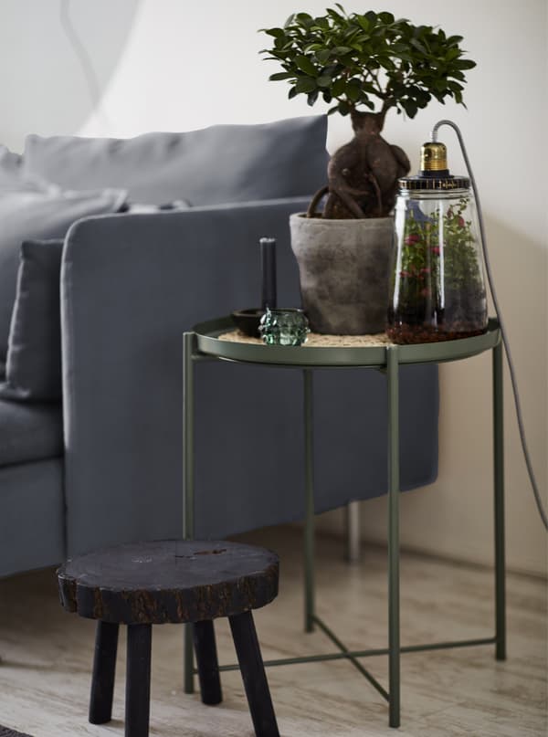 Plante en pot sur une table-plateau à côté d’un canapé et d’un petit tabouret noir.