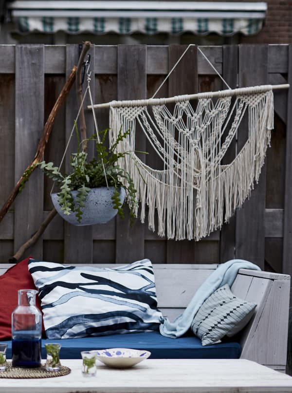 Plantes et macramé accrochés à une barrière, derrière un banc en bois décoré de coussins.