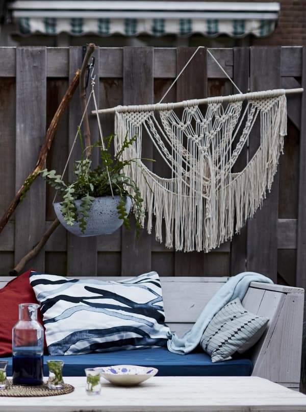 Plants and macramé hanging on a fence behind a wooden bench with cushions.