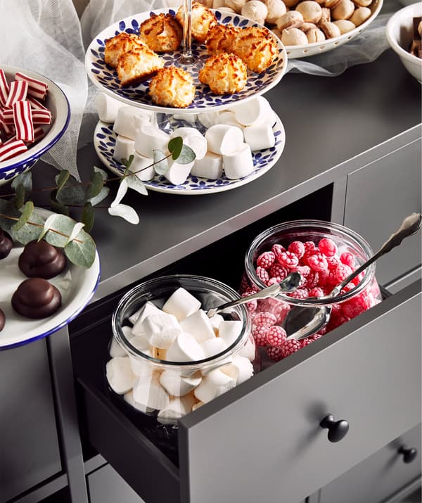 Plats et verres remplis de friandises et de biscuits déposés sur une commode grise. Les tiroirs sont aussi mis à contribution.
