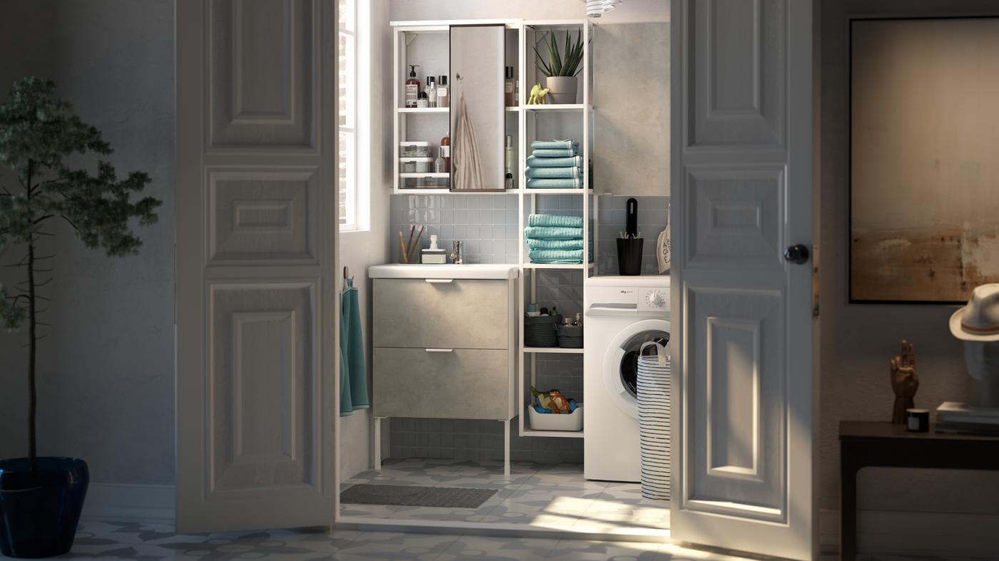 Porte bianche aperte su un piccolo bagno con uno stretto scaffale bianco tra un lavabo bianco e una lavatrice.