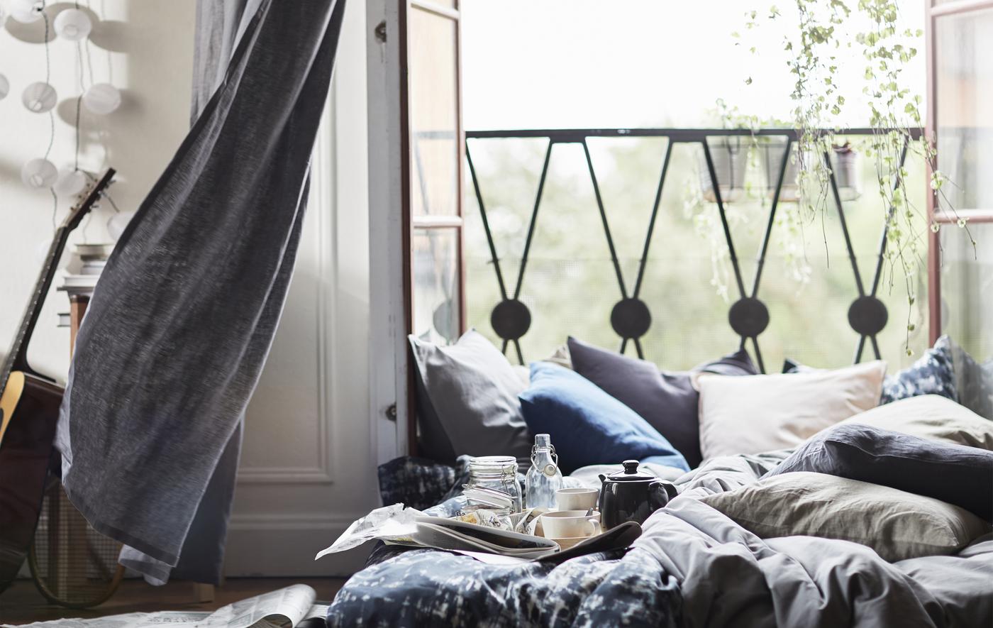 Portes-fenêtres ouvertes avec coussins et literie empilés sur un matelas posé à même le sol.