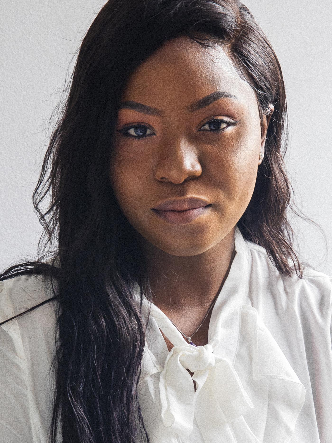 Portrait of Precious, a Nigerian refugee and IKEA Canada co-worker, staring at the camera in a white shirt.