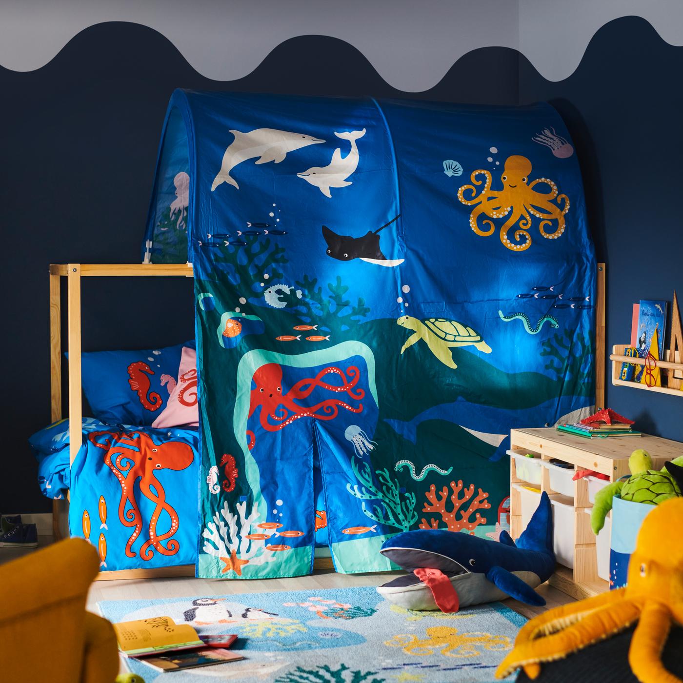 Postel je zakrytá stanem KURA s podmořskými živočichy; modrá plyšová velryba BLÅVINGAD je na koberci BLÅVINGAD před ní.