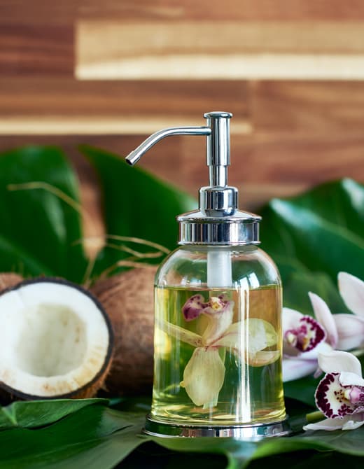 Pot en verre contenant de l’huile à massage et une jolie orchidée ouverte. Plantes tropicales, fleurs et noix de coco complètent le décor. 