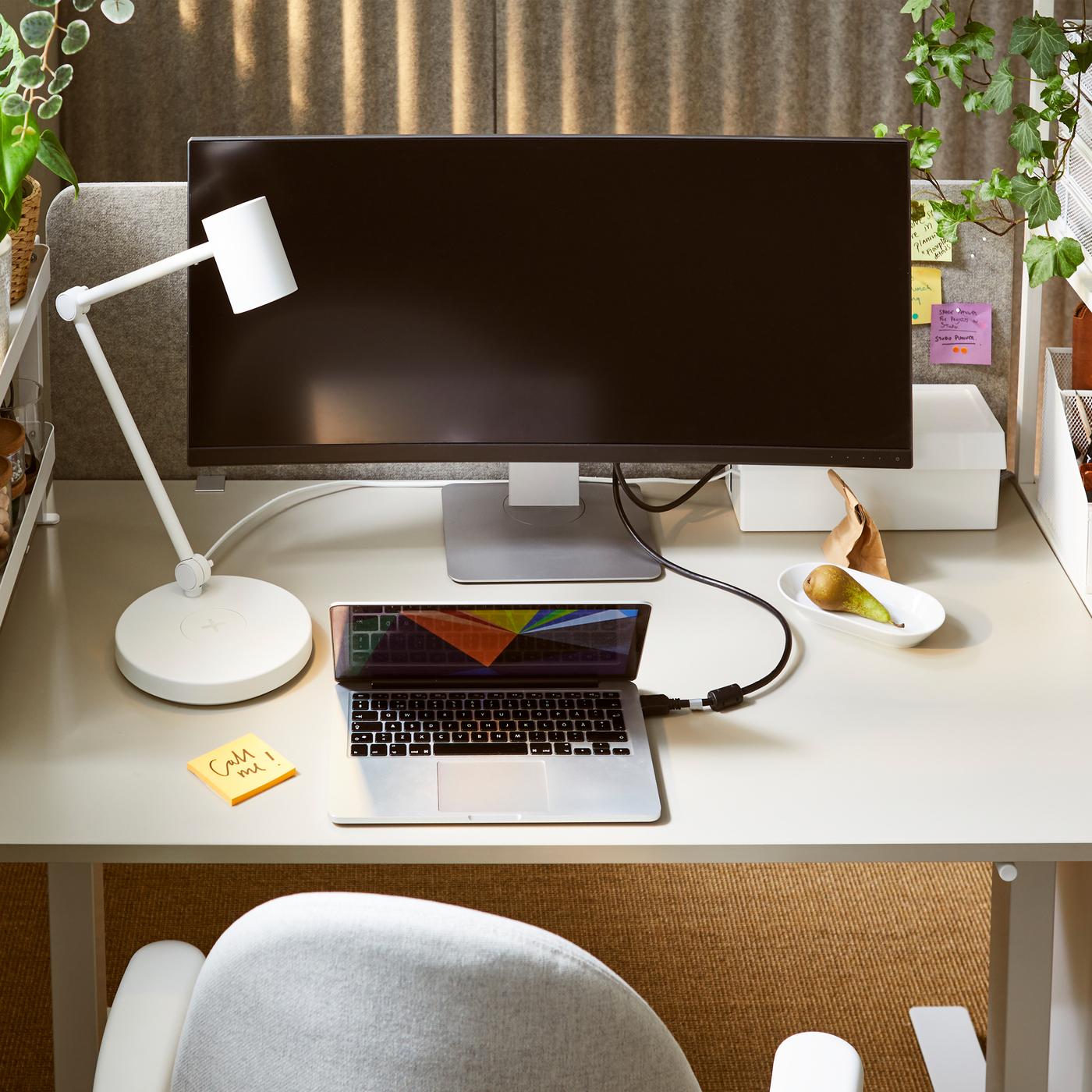Primo piano di una scrivania bianca con una lampada da lavoro NYMÅNE bianca, un ampio monitor, un laptop e una sedia da ufficio grigio chiaro.