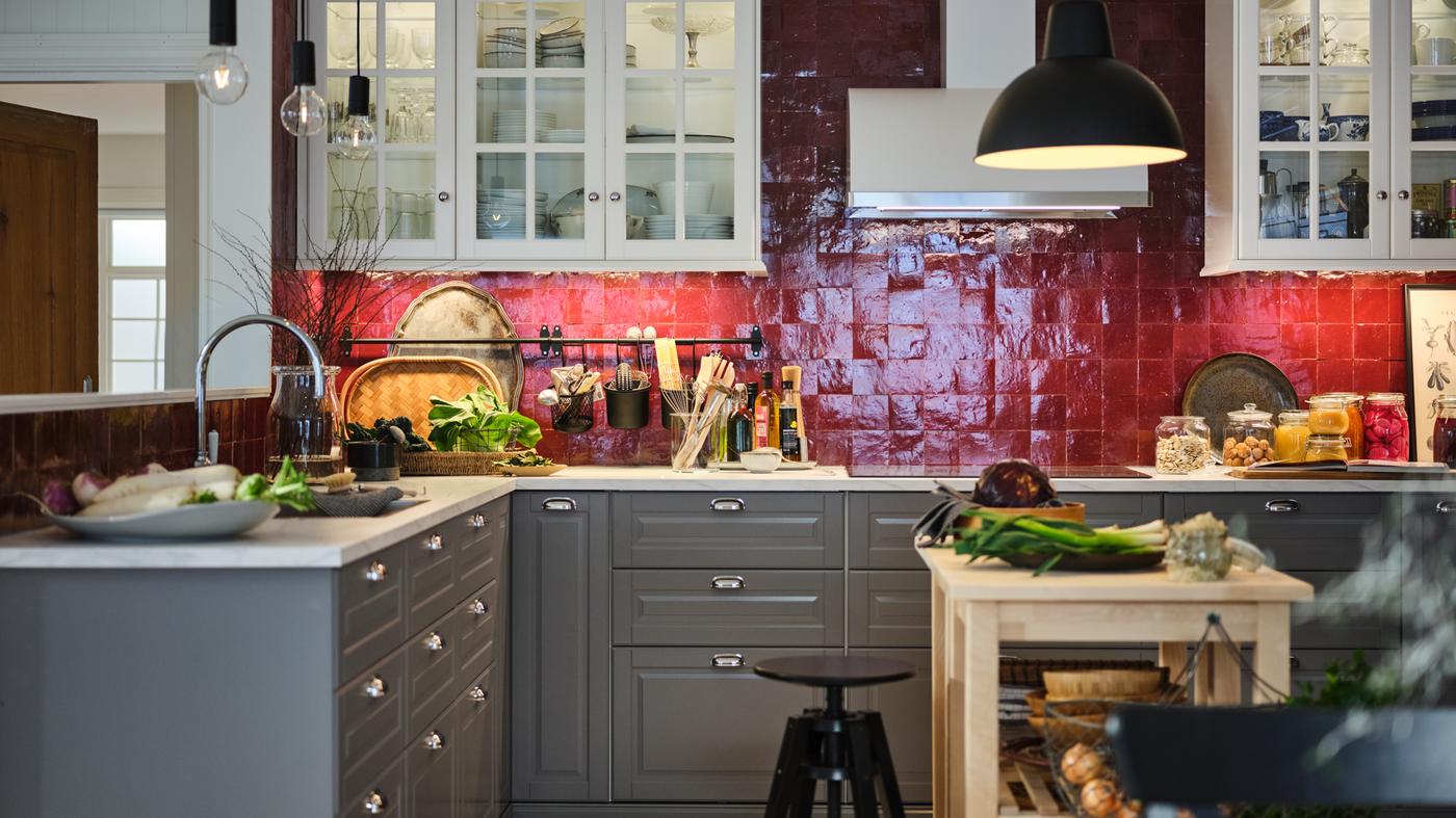 Produce and utensils dot a red-tiled kitchen with white wall cabinets, a white marble effect worktop, and grey base cabinets.