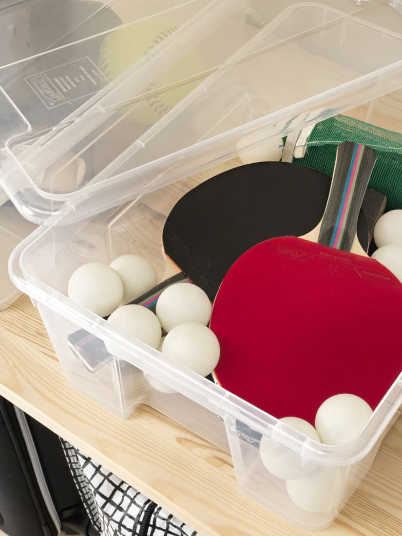Průhledný plastový úložný box IKEA SAMLA s víkem, naplněný vybavením na stolní tenis: červené a černé pálky, bílé míčky a zelený síťový set. Box stojí na dřevěné polici.