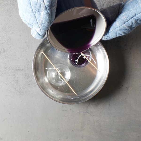 Purple melted wax is carefully poured into glass containers with fixed wicks, as if an IKEA-style DIY candle project is in full swing.