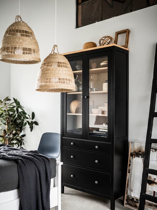 قسم من غرفة نوم فاتحة بها خزانة HEMNES طويلة سوداء ذات أبواب زجاجية، ومصابيح معلقة TORARED، وقطع زينة وديكور، ونبات.