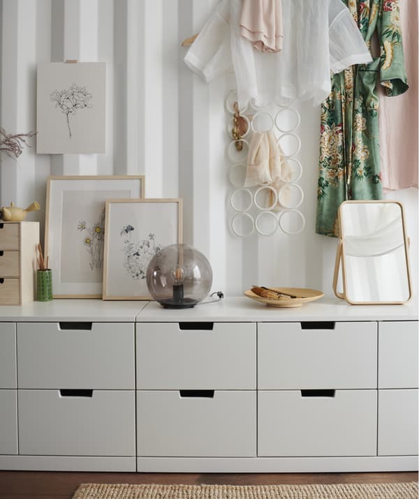 Quadri, una lampada e uno specchio sul ripiano di una cassettiera bianca bassa e vestiti appesi a una parete bianca – IKEA