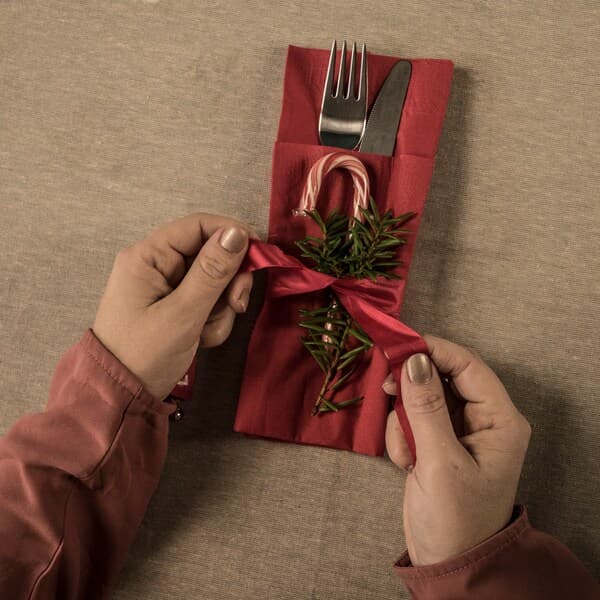 Qualcuno decora un sacchetto porta tovaglioli contenente un coltello e una forchetta con un fiocco, un bastoncino di zucchero e un ramo di abete.