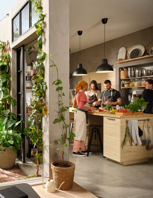 Quatre personnes s’affairant autour d’un grand îlot de cuisine pour la préparation d’un repas. Des portes sont entrebâillées sur une cour agrémentée de plantes.