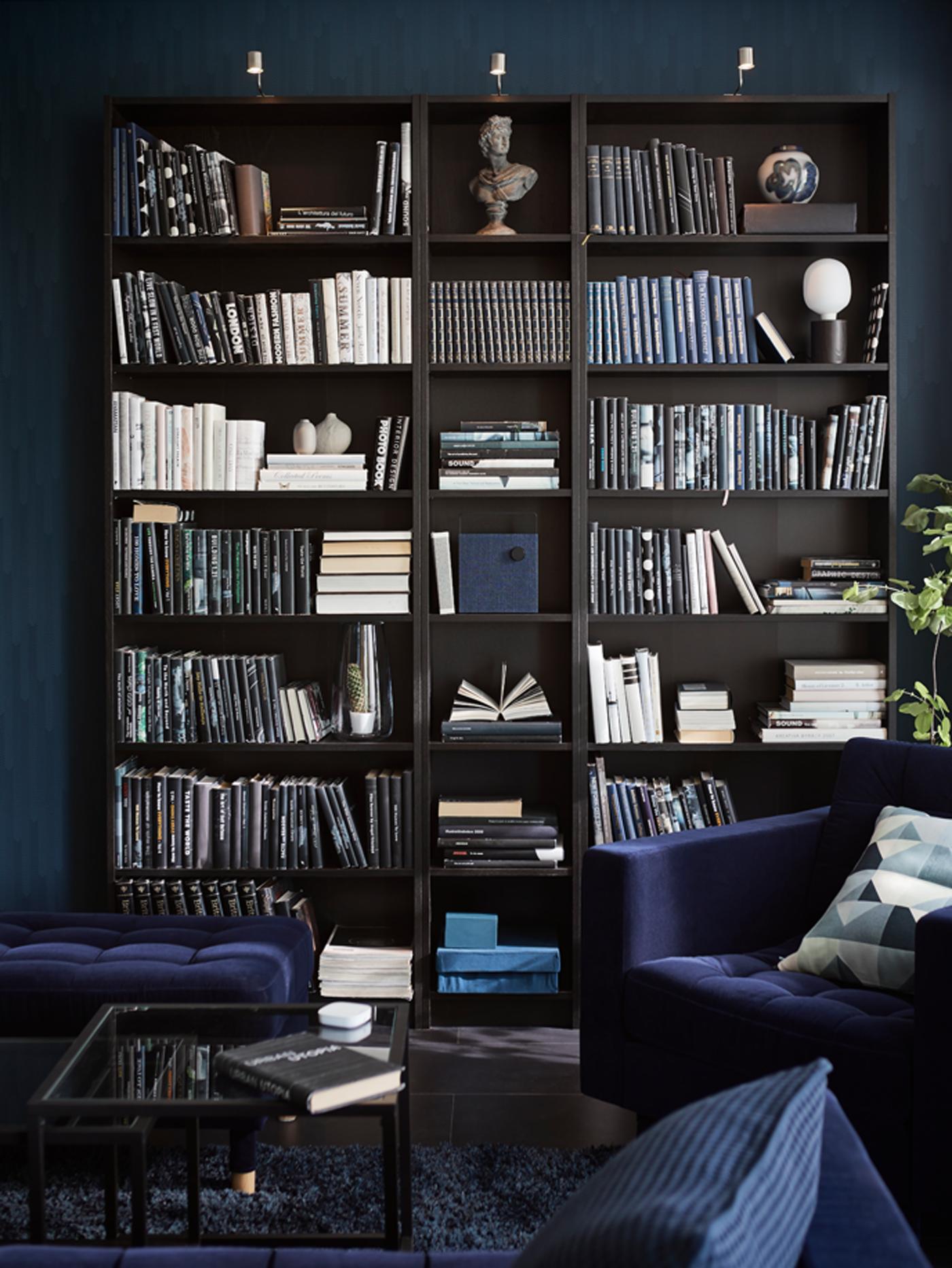 Rak buku BILLY berwarna hitam-coklat yang penuh dengan pelbagai buku dan beberapa objek terletak di ruang tamu berwarna biru. Kerusi berlengan berwarna biru terletak di hadapan.