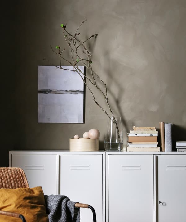 Rangée d’armoires blanches devant un mur gris Sur les armoires, des livres, des articles décoratifs ainsi qu’un vase avec une longue tige et des pousses vertes.