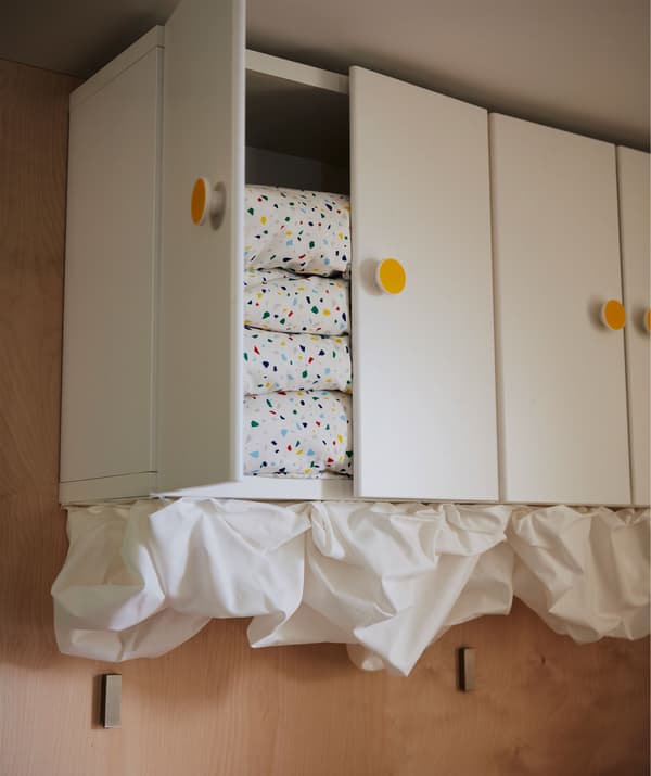 Rangée d’armoires murales touchant le plafond. Couettes empilées dans l’une d’elle. Formes rappelant les  nuages créées avec une pièce de tissu.
