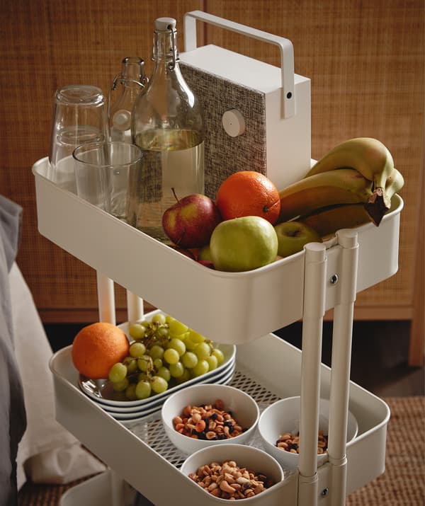 RÅSKOG trolley filled with various snacks and fruits, a KORKEN bottle and drinking glasses, and an ENEBY Bluetooth speaker.