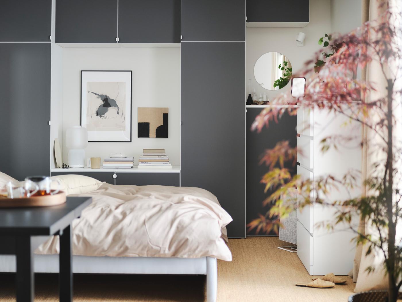 Recámara con una cama con ropa de cama beige, una combinación de clósets grises de fondo, una planta y una mesa negra en primer plano.