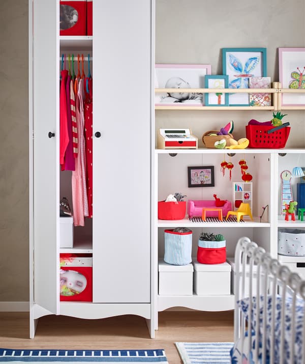 Red clothes and boxes in a tall white wardrobe, and books, toys, and boxes on open shelves.