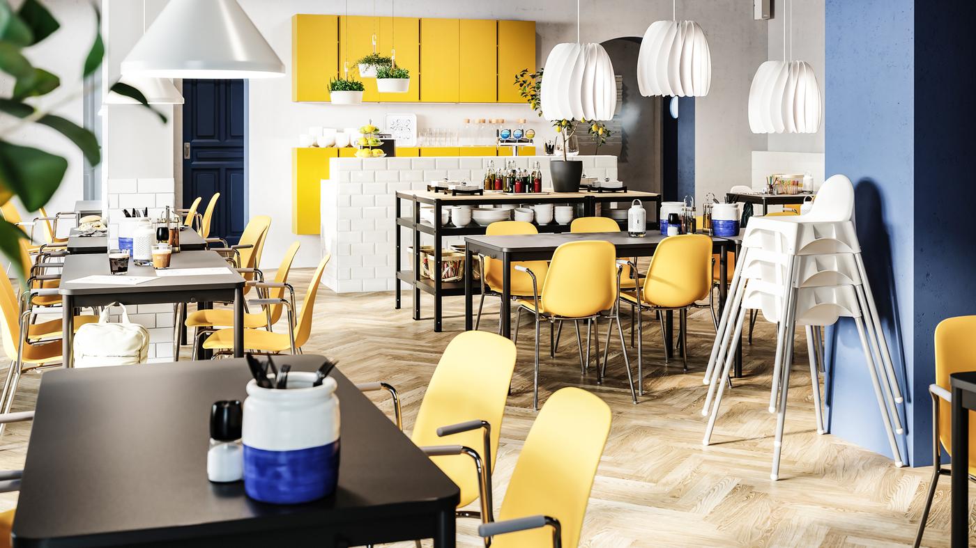 Restoran yang cerah dengan kerusi berwarna kuning gelap, meja berwarna hitam, kerusi junior berwarna putih, kabinet berwarna kuning dan lampu pendan berwarna putih.