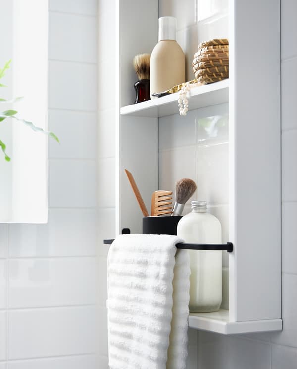 رف حائطي HEMNES أبيض عليه زجاجات وماكياج وفوطة بيضاء على علّاقة معدنية سوداء مثبتة على الرف.