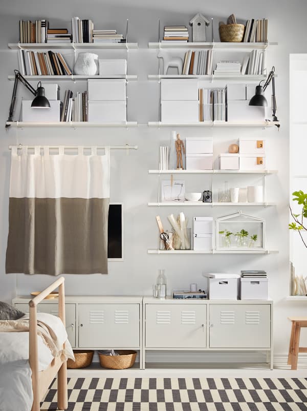 رفوف BERGSHULT/PERSHULT بيضاء مملوءة بالتخزين والديكورات، وخزائن IKEA PS بيضاء تغطي جدار غرفة النوم.