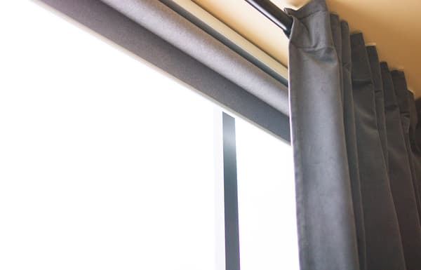 Rolled-up FYRTUR smart blind in grey in front of a window, and behind a grey curtain.