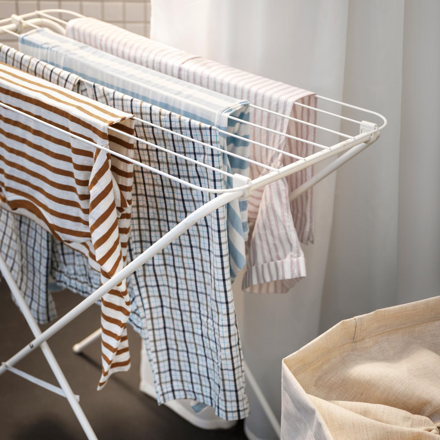 Ropa a rayas y cuadros colgada en un tendedero JÄLL blanco frente a una cortina de baño blanca, detrás de una bolsa de lavandería beige.