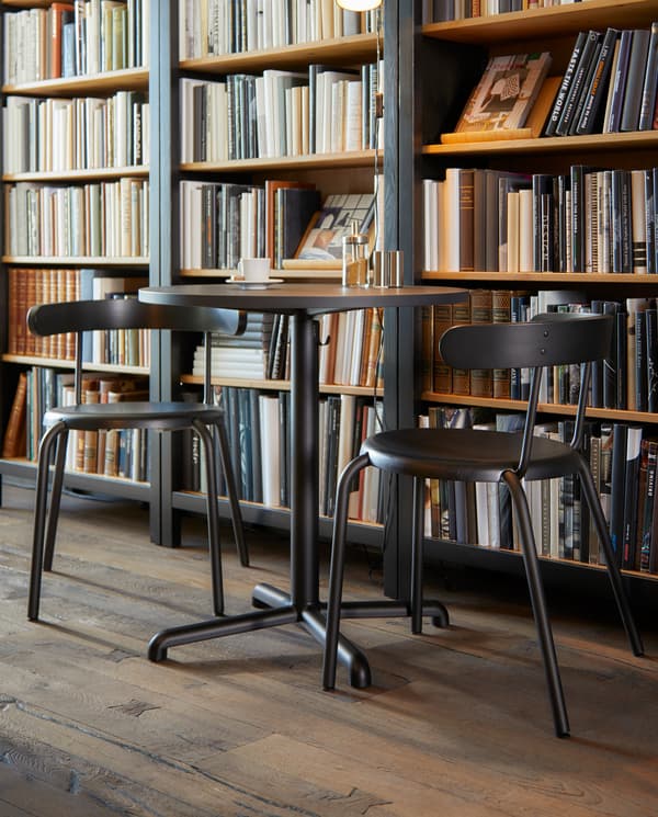 Round anthracite café table with hooks for bags underneath, two chairs, in front of a large bookcase filled with books.