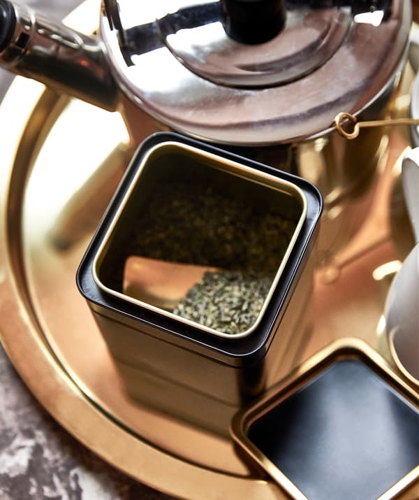 Round, golden tray with tea making kit on it: kettle, a tin with its lid off revealing green tea inside, cups on the side.