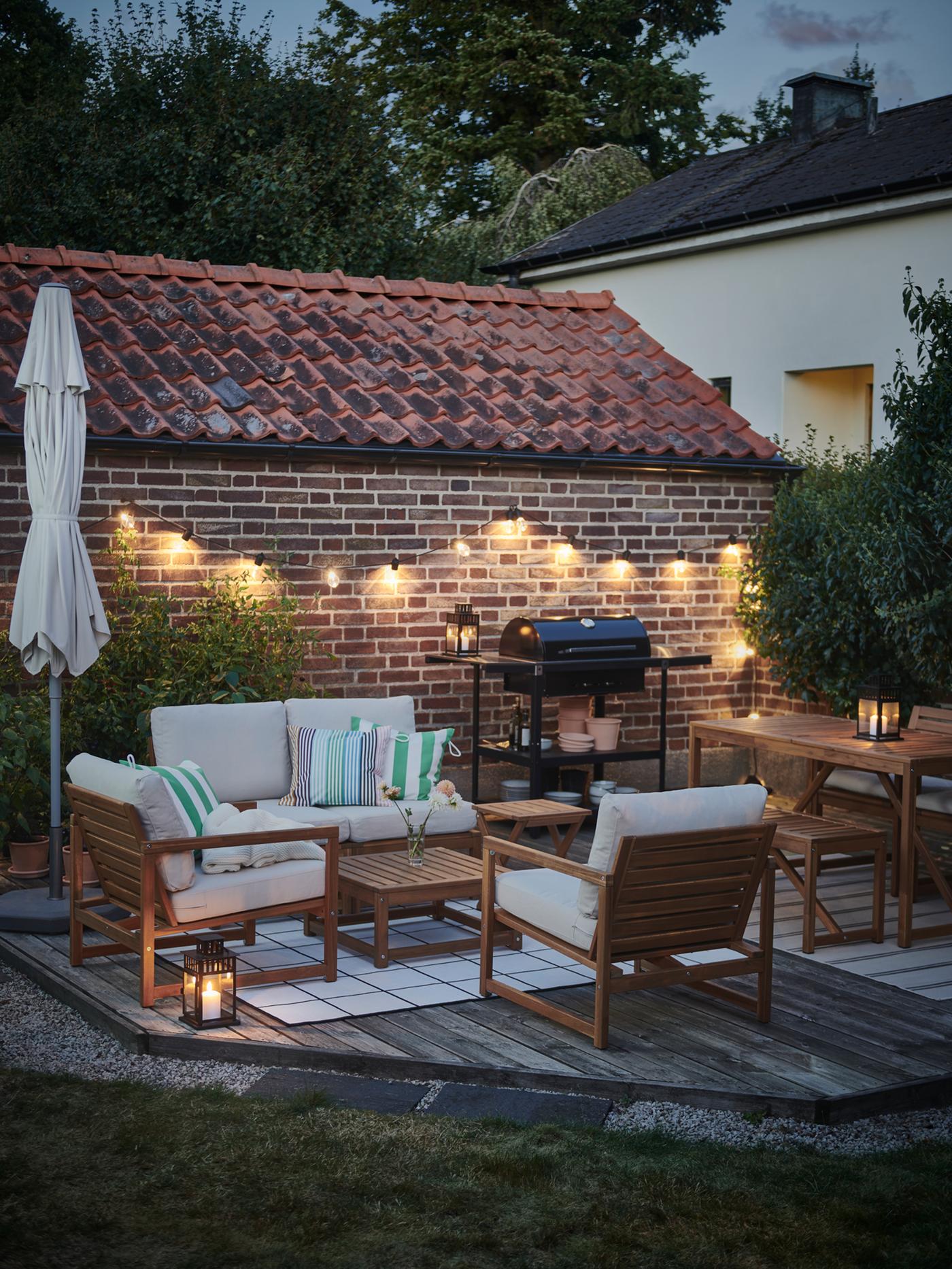 Ruang luar senja kala dengan set santai empat tempat duduk NÄMMARÖ, barbeku GRILLSKÄR dan rantaian cahaya LED SVARTRÅ.