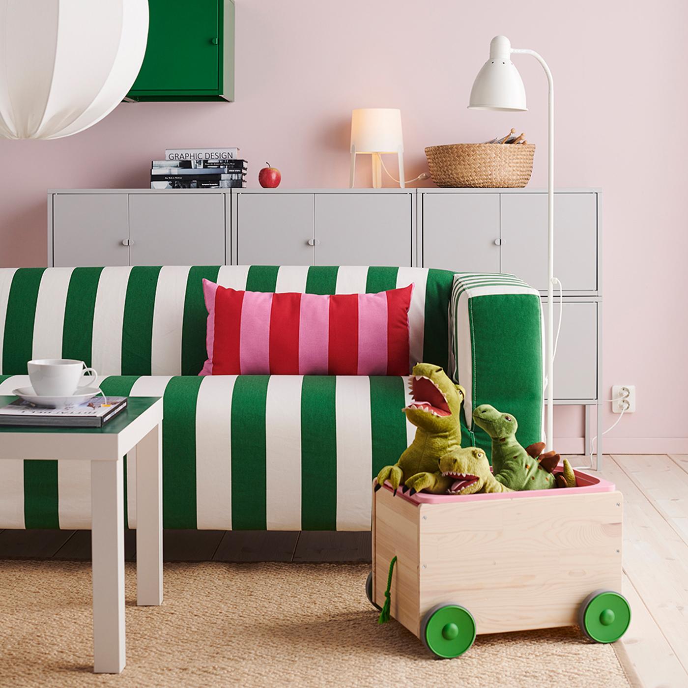 Ruang tamu yang mempunyai sofa KLIPPAN, beberapa kabinet LIXHULT berwarna kelabu, meja LACK berwarna putih dan troli yang penuh dengan dinosaur mainan lembut.