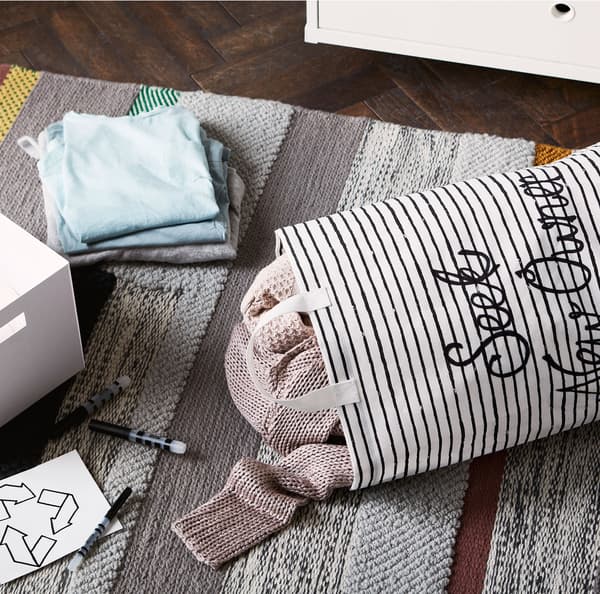 Sacco per il bucato KLUNKA pieno di maglie, una scatola bianca e una pila di vestiti sparsi su un tappeto grigio - IKEA