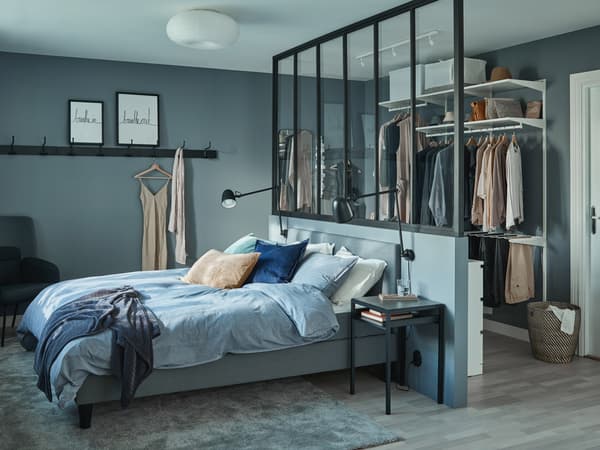 SÄBÖVIK bed in a bedroom draped with blue beddings, resting against a room divider. A closet hanging at the back of the room.