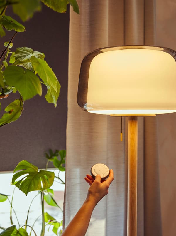 Šaka drži TRÅDFRI daljinski upravljač pored upaljene EVEDAL podne lampe. Iza se nalazi prostor, delimično pokriven TRETUR roletnom.