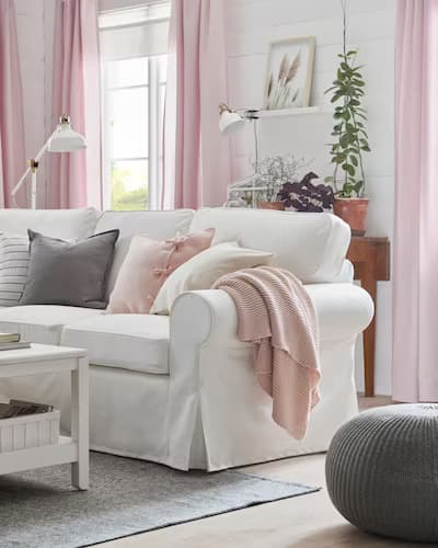 Sala con sillón blanco decorado con cojines y mantas en tonos grises y rosados. Además hay cortinas rosadas y una mesa de centro de color blanco.