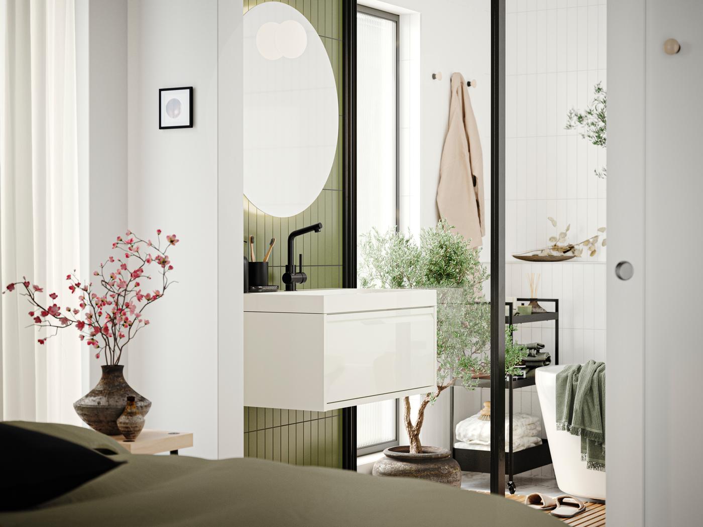 Salle de bain avec meuble lavabo à tiroir ÄNGSJÖN en blanc brillant, miroir et baignoire vus d’une chambre.