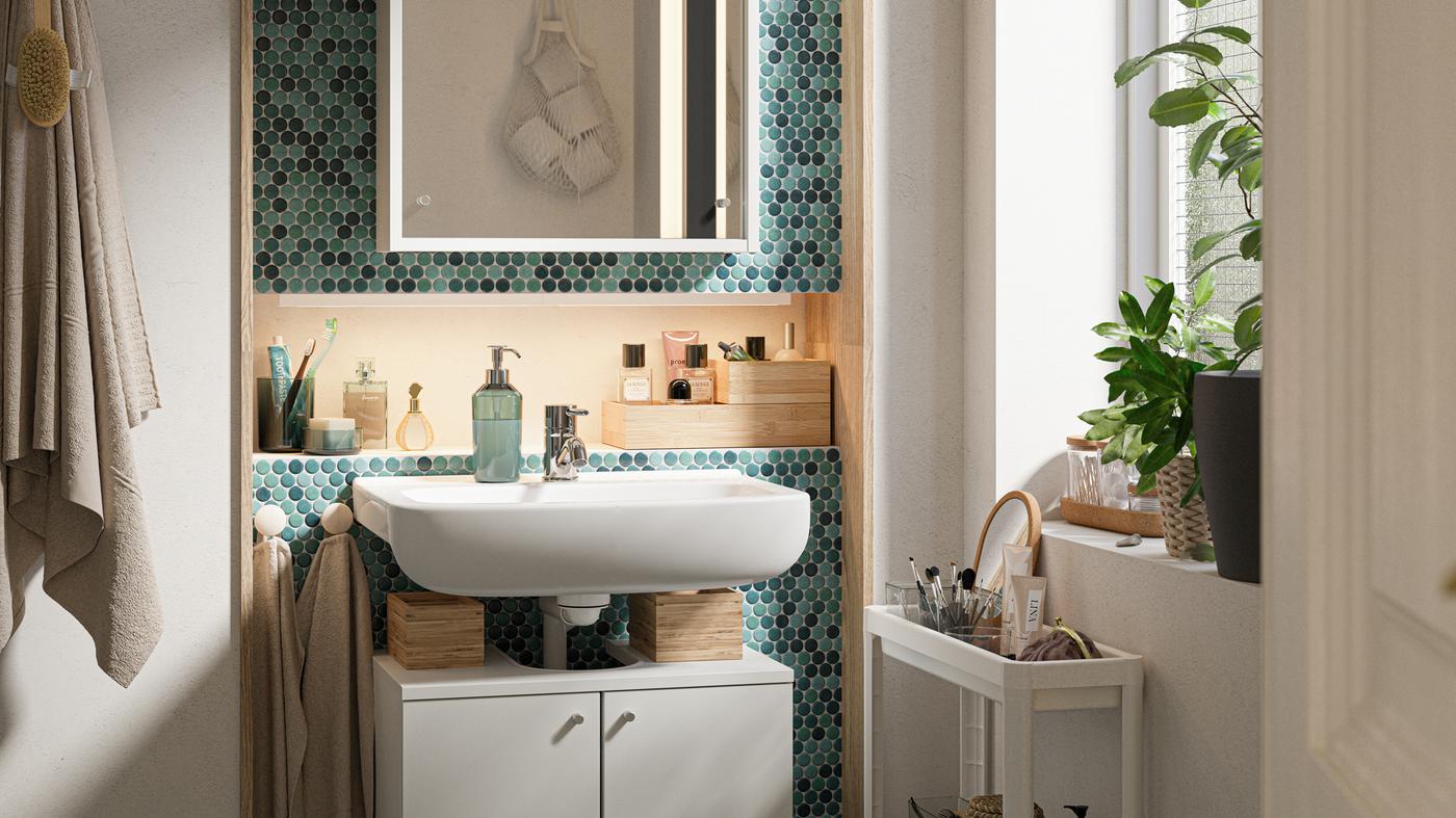 Salle de bain carrelée de vert avec mobilier blanc, plantes en pot et desserte blanche chargée d’articles de toilette.