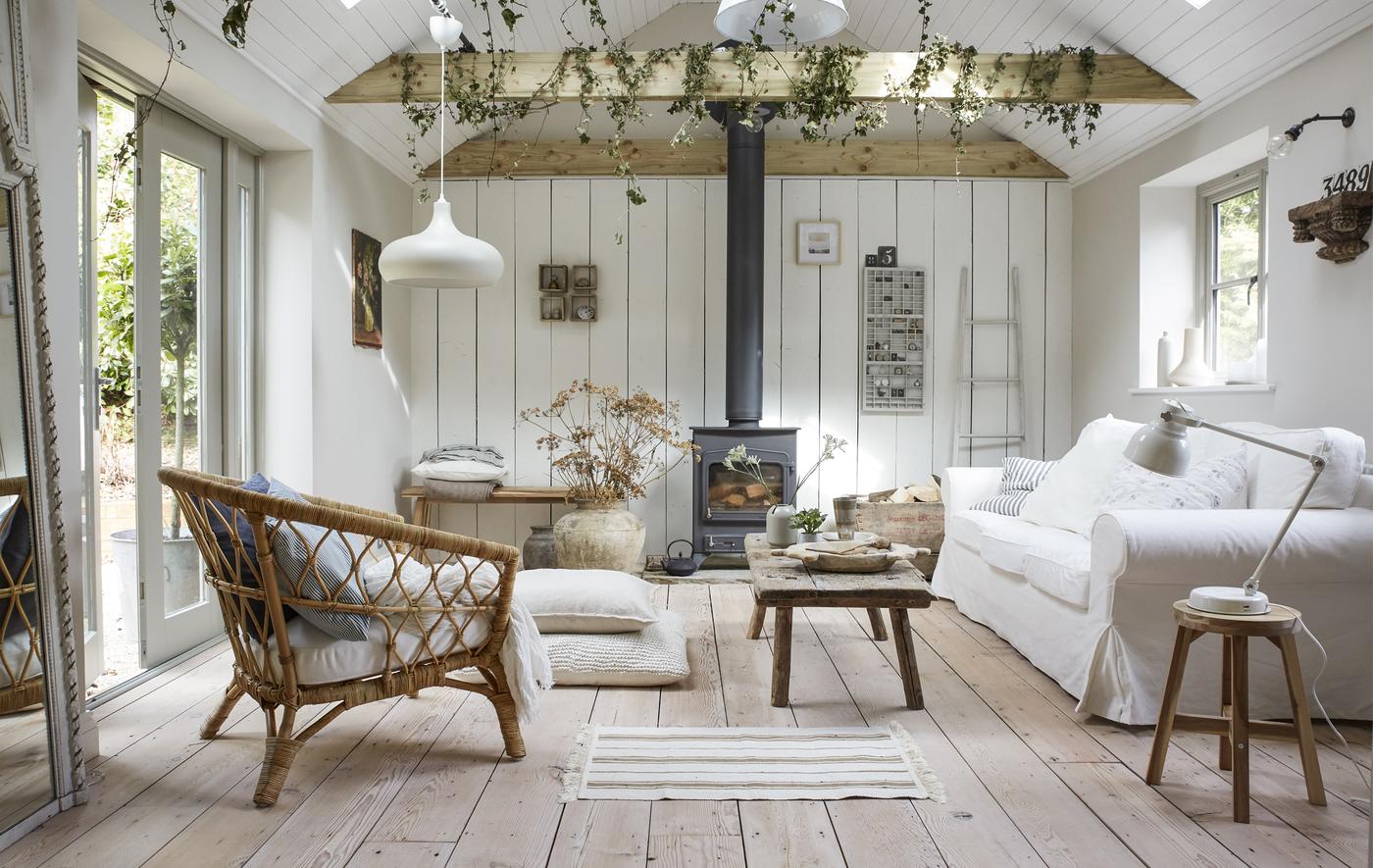 Salon de couleur claire décoré d’un canapé blanc et d’une chaise en osier, avec poutres et planchers en bois.