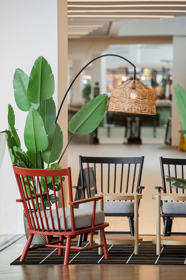 Scandinavian-style seating area with red, black, and natural wood armchairs, gray cushions, a woven rattan floor lamp, artificial green plants, and a black grid-pattern rug in a modern showroom setting.