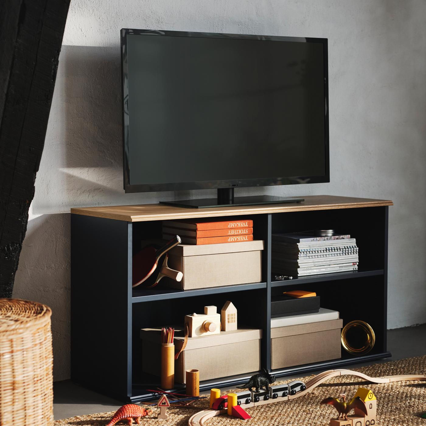 Scatole, libri e giocattoli riempiono i ripiani a giorno di un mobile TV SKRUVBY nero-blu, su cui è posizionata una TV nera.