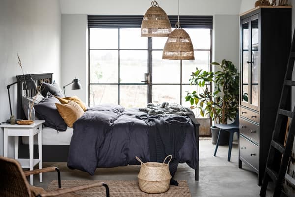 Schlafzimmer mit einem dunklen HEMNES Bettgestell in der Mitte, Flügeltüren entlang der Wand und Details aus Naturmaterialien bzw. in Naturfarben.