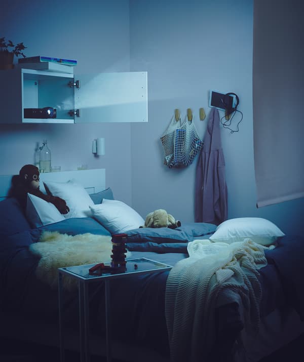 Schwach erhelltes Schlafzimmer, in dem u. a. ein BESTÅ Regal mit Tür zu sehen ist, in dem ein Beamer steht. 