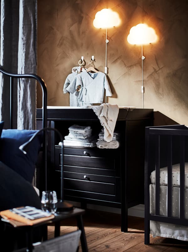 Section d’une chambre, à moitié dissimulée derrière un rideau séparateur, où se trouvent une table à langer et un lit à barreaux SUNDVIK noir-brun.