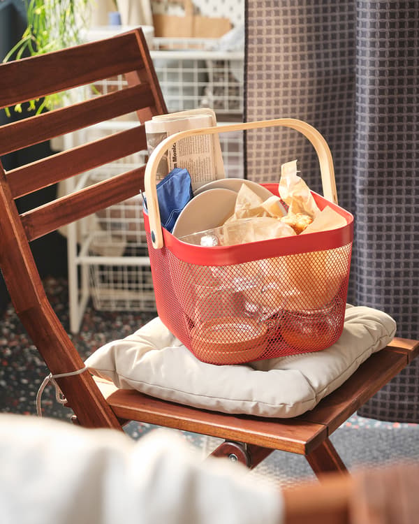 Section of balcony with red RISATORP basket packed with a small meal, placed on the seat cushion of an ÄPPLARÖ chair.