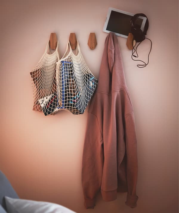 Section of bedroom wall fitted with row of SKUGGIS bamboo hooks holding filled net bags, a hoodie, and a tablet with earphones.