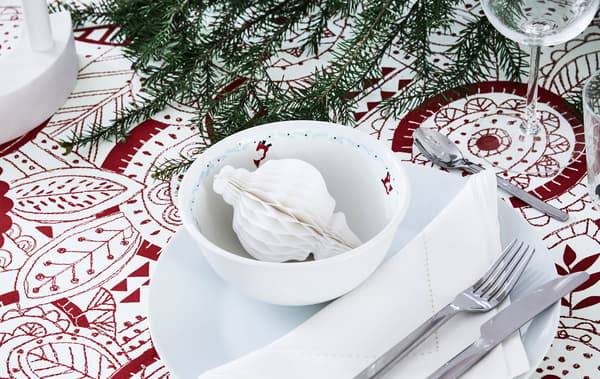 Section of dinner table set for a festive occasion. Patterned tablecloth, LED candle, spruce twig and paper decoration.