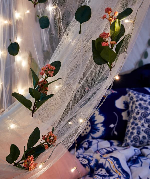 Section of mosquito net draped over a bed at night, the net decorated with artificial flowers and multiple lighting chains.