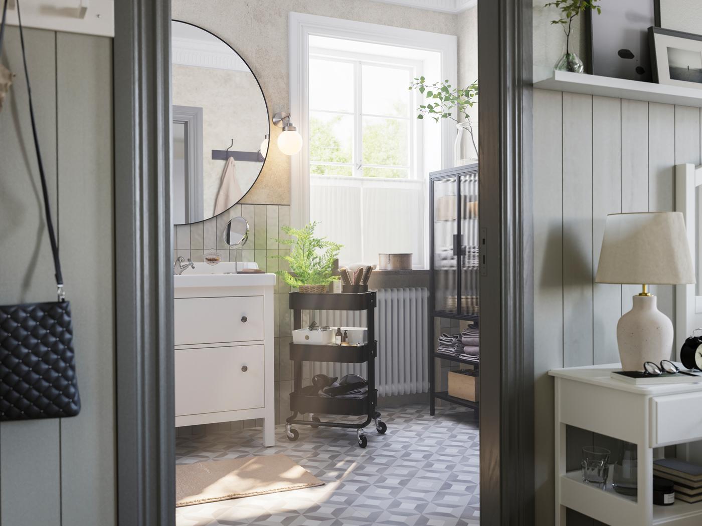 Šedá koupelna s velkým oknem. Je zde bílý umyvadlový stolek HEMNES, černý vozík RÅSKOG a skleněná skříňka MOSSJÖN.